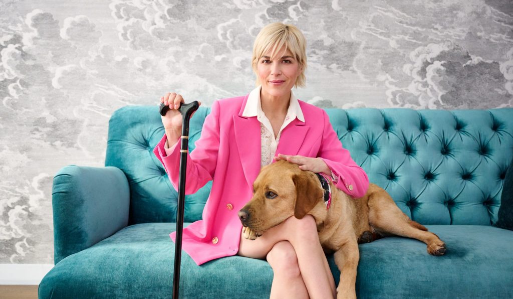 Selma Blair, QVC Brand Ambassador for Accessibility, sits on a plush teal sofa with her dog, Scout. She wears a hot pink suit and uses a black and gold cane.