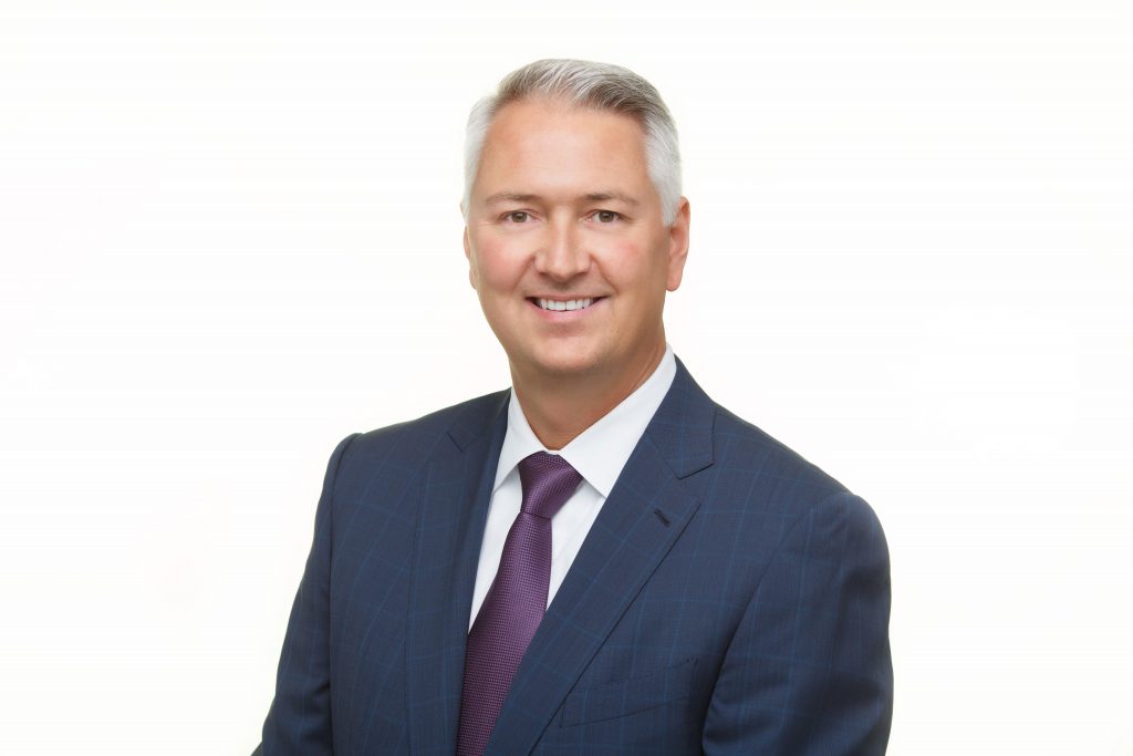 A man in a business suit, with a blue windowpane jacket, white shirt and purple tie.