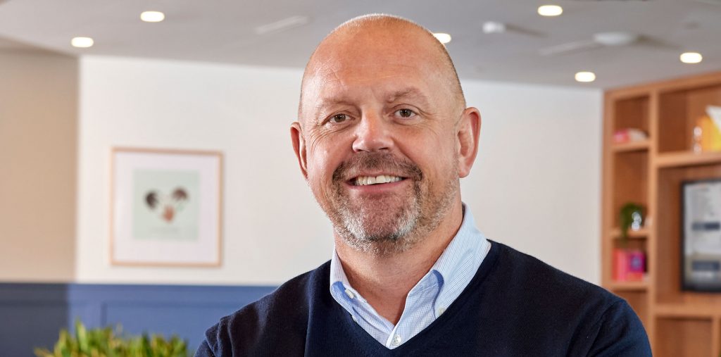 Mike Fitzharris standing in an office, wearing a light blue shirt with an open collar and a dark blue sweater.